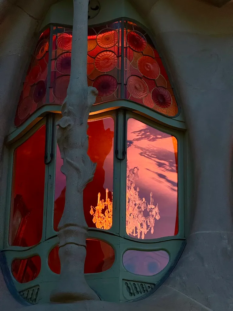 Close-up of an ornate Modernist window with a bone-like stone frame, characteristic of Gaudí's Casa Batlló. The upper portion features circular stained-glass panels in vibrant reds, oranges, and purples resembling swirled discs. Through the lower glass panes, a crystal chandelier with lit candle-style bulbs is visible, casting a warm glow against a red-orange interior.