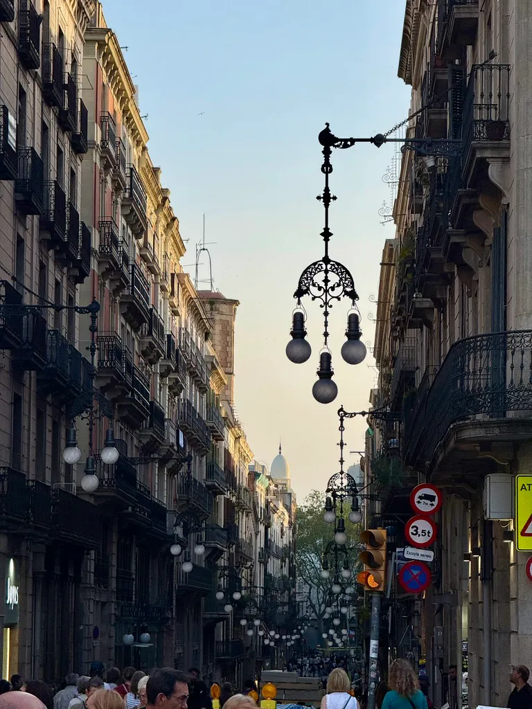 View down a narrow Barcelona street lined with historic multi-story buildings featuring wrought-iron balconies. An ornate black cast-iron street lamp with four globe lights hangs prominently in the center, suspended between buildings. Similar decorative lamps recede into the distance, where a domed building is visible against a pale evening sky. Pedestrians walk along the street below, and traffic signs are mounted on the right.