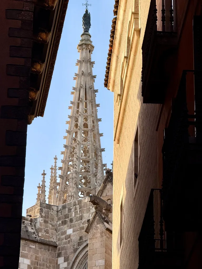 Looking up between two buildings at the intricate Gothic spire of Barcelona Cathedral. The tall, tapering stone tower features elaborate openwork tracery, pinnacles, and a statue topped with a cross at its peak. A gargoyle juts out from the cathedral's stonework in the mid-ground. The scene is framed on the left by a dark brick building and on the right by a smooth ochre-colored wall with a balcony, set against a clear blue sky.