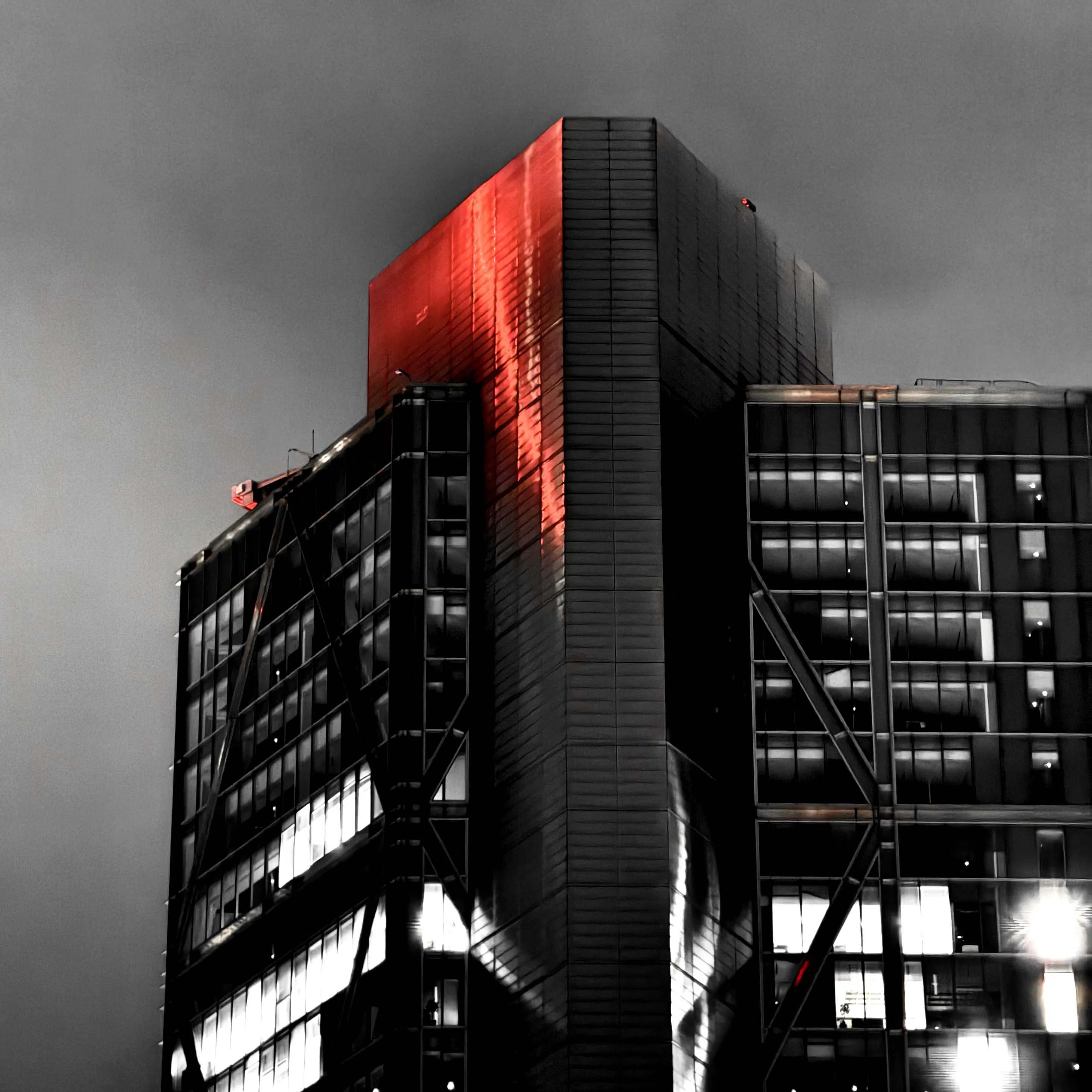 A high contrast black-and-white photo of a highrise building against a monotone dark-grey and cloudy background. Sharp red light illuminates the top of the building.